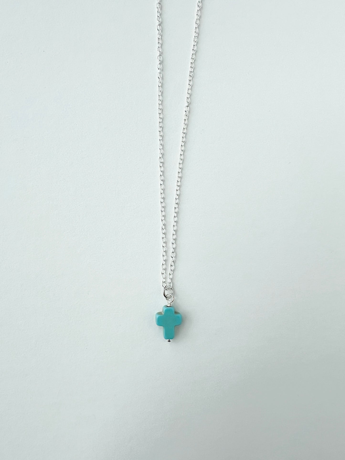 Turquoise cross charm pendant on a silver chain against a light gray background