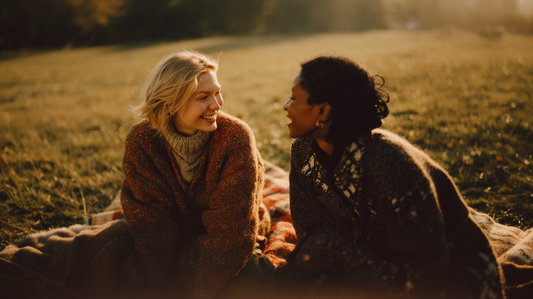 Two young women, one with blonde hair and fair skin and one with dark hair and dark skin, wearing big sweaters and looking at each smiling. The women are sitting on a blanket on a fall day in the sunshine.