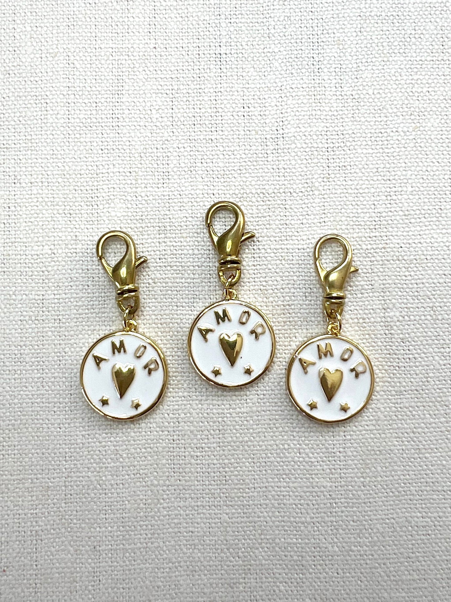 Three gold and white charms with 'AMOR' text on a light gray background