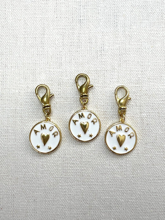 Three gold and white charms with 'AMOR' text on a light gray background