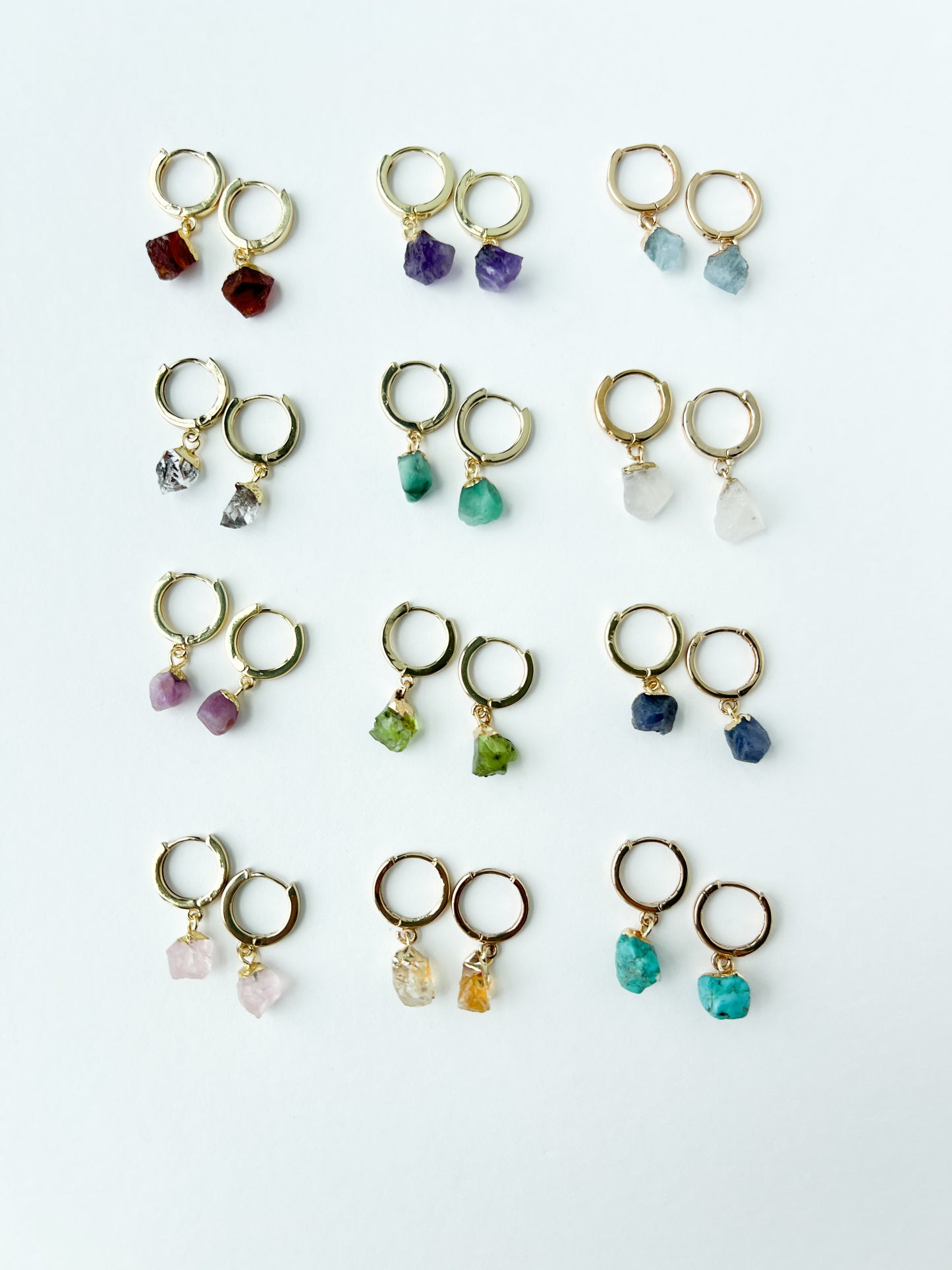 Collection of gold earrings with various colored raw gemstone birtstones on a white background