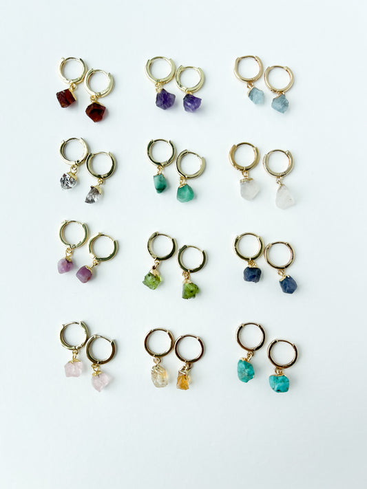 Collection of gold earrings with various colored raw gemstone birtstones on a white background