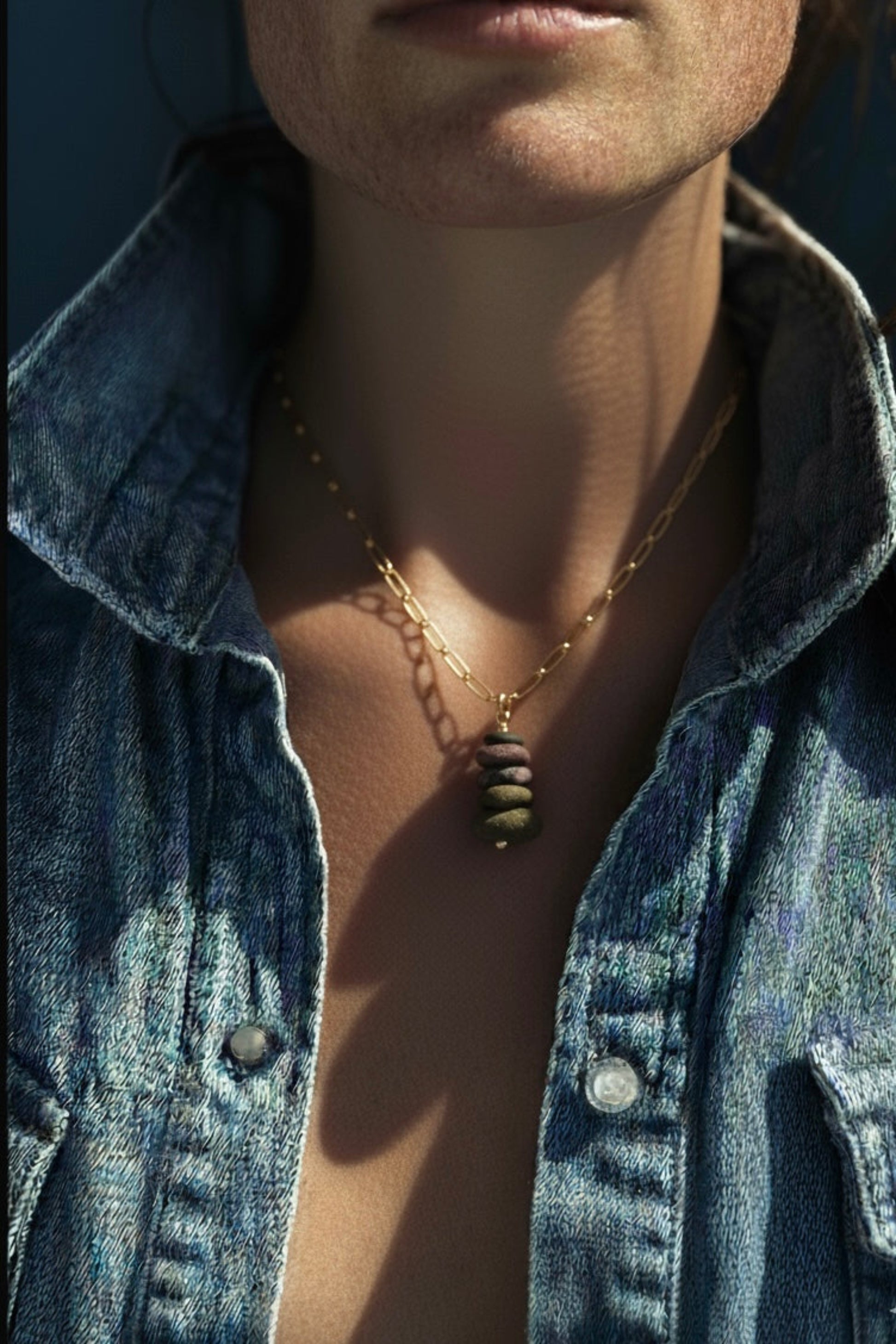 A woman in a denim shirt with collar up wearing a gold paper clip chain necklace with a stacked Michigan lake stone charm.