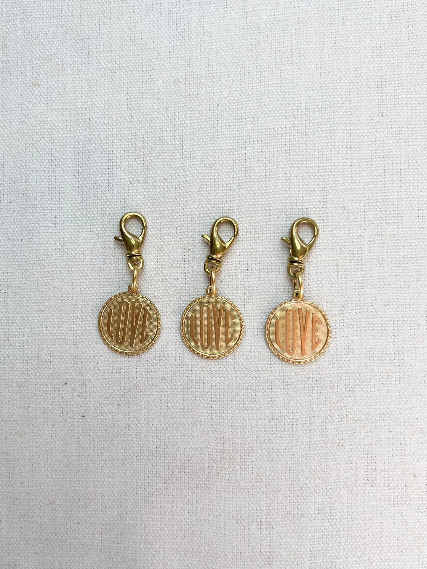 Three gold charms with 'LOVE' engraving on a light fabric background