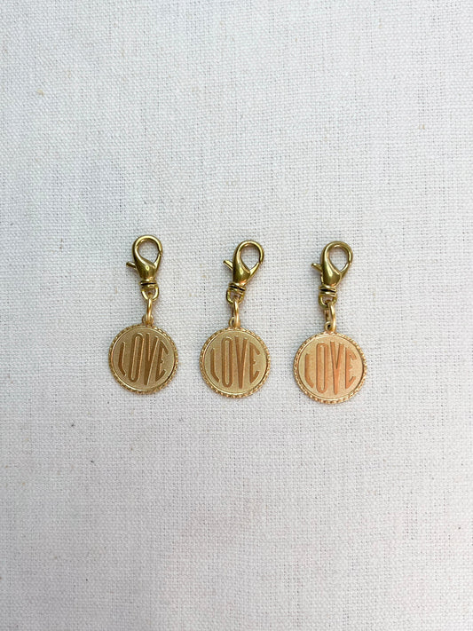 Three gold charms with 'LOVE' engraving on a light fabric background