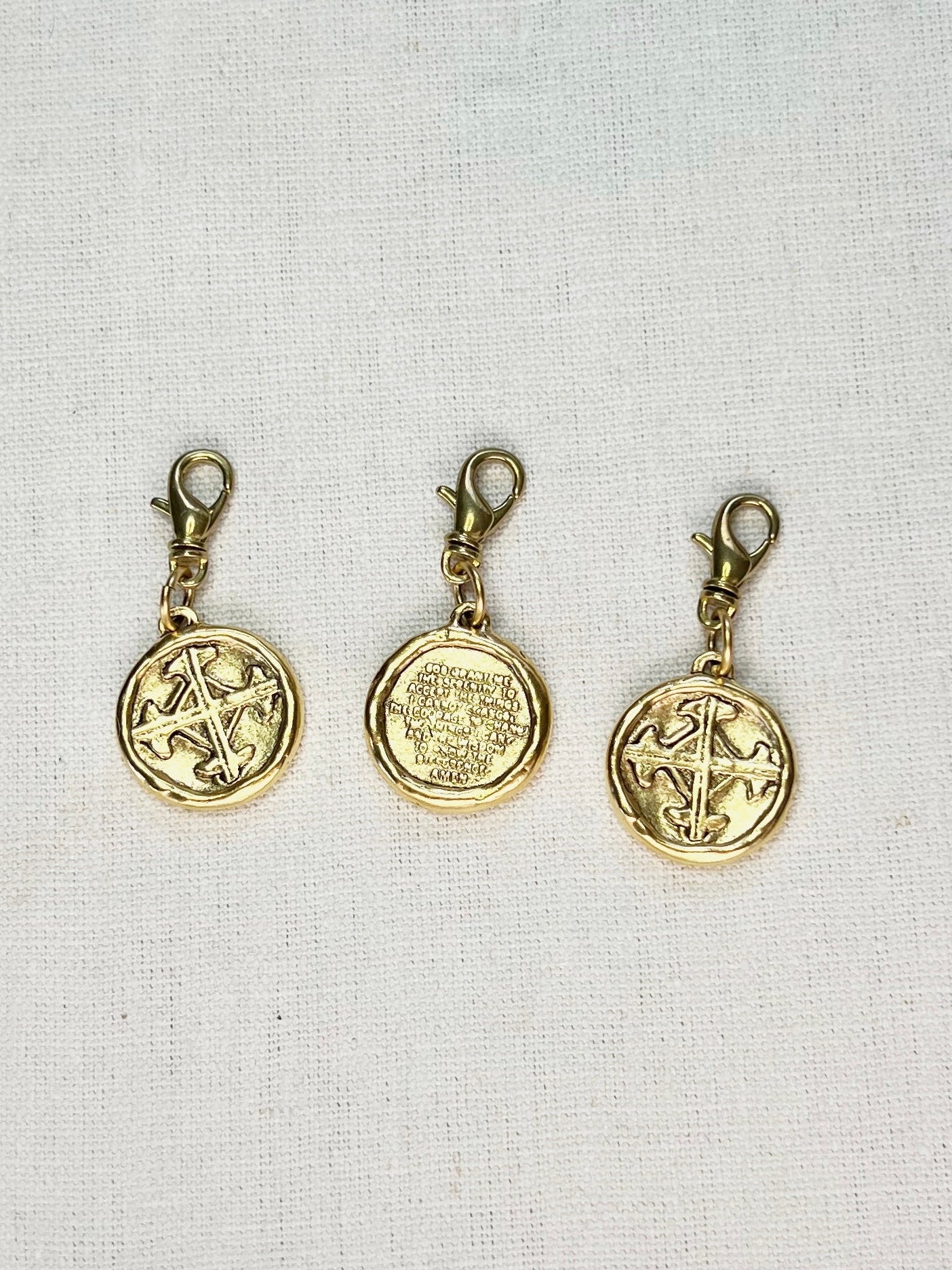 Three gold coin-shaped charms with the serenity prayer and lobster clasps on a light fabric background