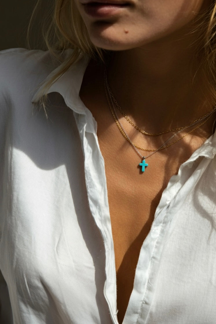 A woman wearing a white button down shirt with a small turquoise cross on a silver chain and two gold chains