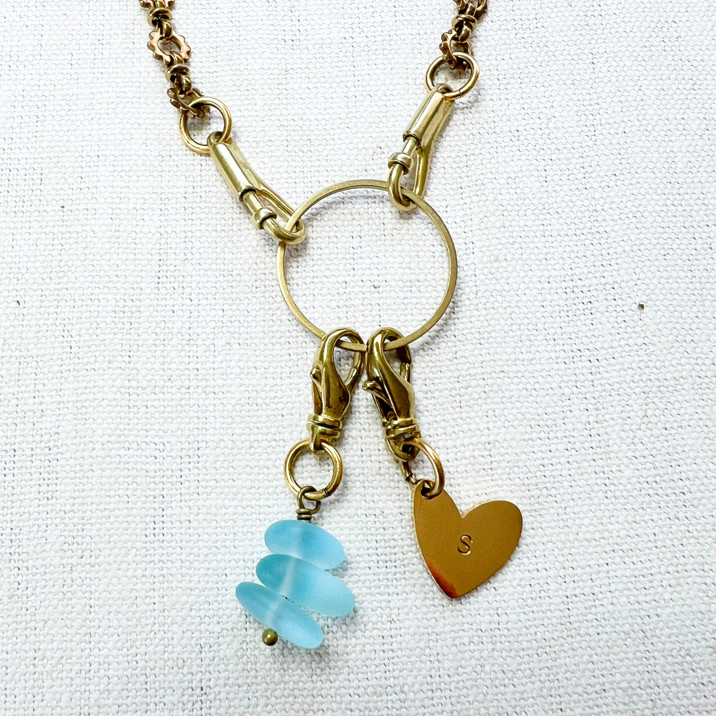 Necklace with gold chain, heart charm, and blue sea glass pendant on a white fabric background